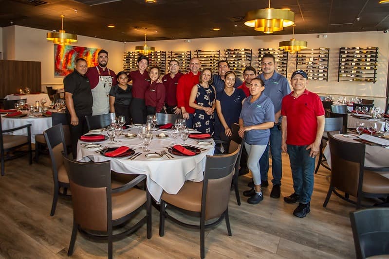 Cibao Grille restaurant team standing together in an elegant dining room with set tables, showcasing professional staff, warm ambiance, and fine dining experience.